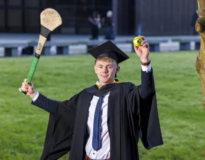 A photo of a man in smart dress and dark graduation robes and cap, holding a hurley in one hand and a sliotar in the other