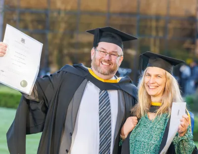 A photo of a man and woman in smart dress and graduation robes and cap, each holding up a degree certificate