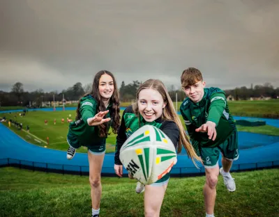 three youth touch rugby athletes, one male and two female, leaning towards camera with a rugby ball with a running track in the background