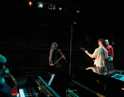 Three performers and musicians on stage while two others sitting next to a piano