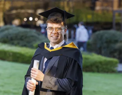 Man standing in outdoor setting wearing graduation robes and holding graduation scroll