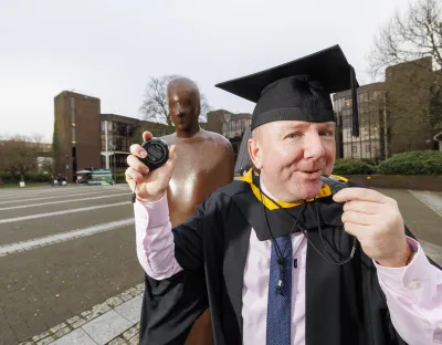 Man in graduation robes holding a coaches whistle and stopwatch standing in front a bronze statue with university building in the background