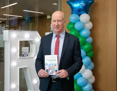 Professor Alan Donnelly posing with the HRI Annual Report in front of a stack of balloons