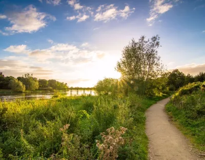 Path on the right of a river with blue skies and green bushes on the left