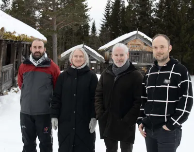 A photo of four people dressed in outdoor clothing, standing in snow in front of cabins and trees