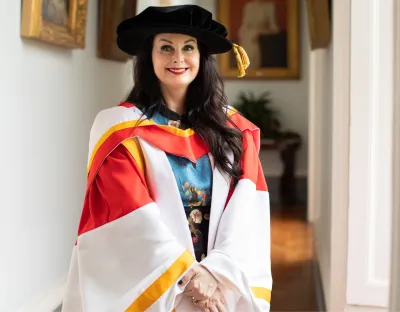 A photo of a woman in red and white conferring robes with a black conferring cap, standing in a bright hallway with art in the background