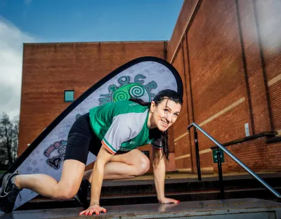 A female athlete with dark hair, wearing a green and white sports jersey, crouches on a metal platform, smiling at the camera. She appears to be mid-obstacle in an outdoor setting, with a brick building and a branded flag in the background. The flag displays a green spiral design with the words "OCR Ireland." The scene is set against a partly cloudy sky.