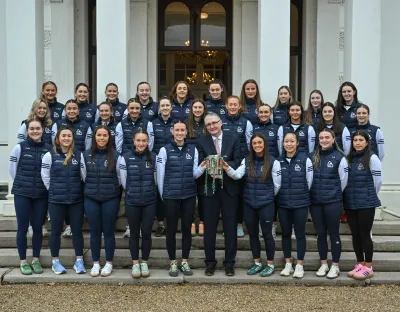 Acting President of the University of Limerick, Professor Shane Kilcommins, stands with the UL Ashbourne Cup-winning camogie team outside Plassey House. The team, dressed in navy UL jackets and white jerseys, proudly gathers around him as he holds the prestigious trophy. Smiling faces reflect their back-to-back championship success for UL camogie.