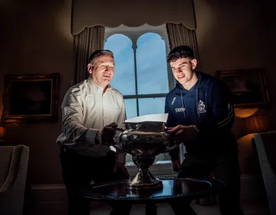 Two men, an older one to the left wearing a light shirt, and a younger man to the right wearing a navy hoody, in a stately setting, holding a champioship cup and looking into the bowl. There is a stately window behind them