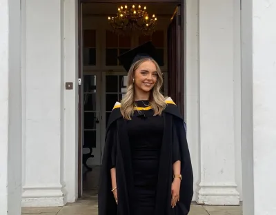 A photo of a woman in black graduate robes and cap, standing in front of a doorway