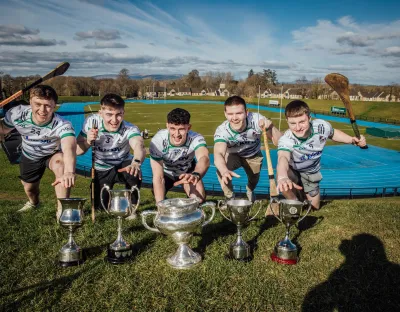 Five young male hurlers in white and green University of Limerick jerseys pose on a grassy hill, reaching toward five silver trophies placed in front of them.  Behind them, the university's blue athletics track and green playing fields are visible under a bright blue sky