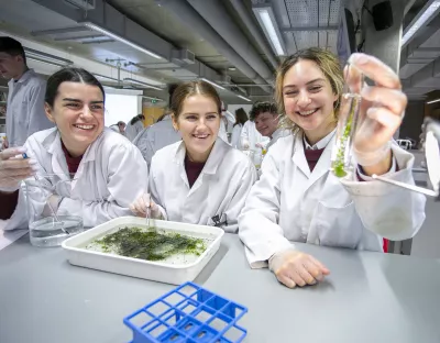 Students in lab coats and glasses smiling while observing a test tube filled with green matter 