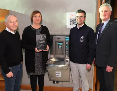 Four people gathered around a water bottle filling facility on campus with one person holding an award