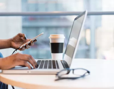 Image depicting a work desk with coffee, glasses, and a laptop. A woman's hand is holding a mobile phone.