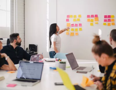 Woman placing sticky notes on a wall as a team of people sitting at a desk with laptops watch her.