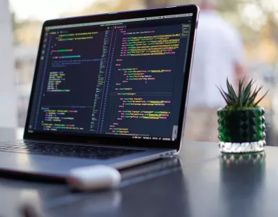 Laptop turned on with code displaying on the screen and a plant next to the laptop in a coffee shop.