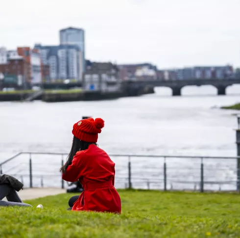 Two students sit on a green wearing winter clothes with Limerick City and the River Shannon in the background