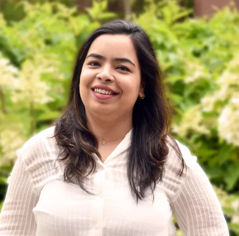 A young woman in a white top standing in front of flowers smiling