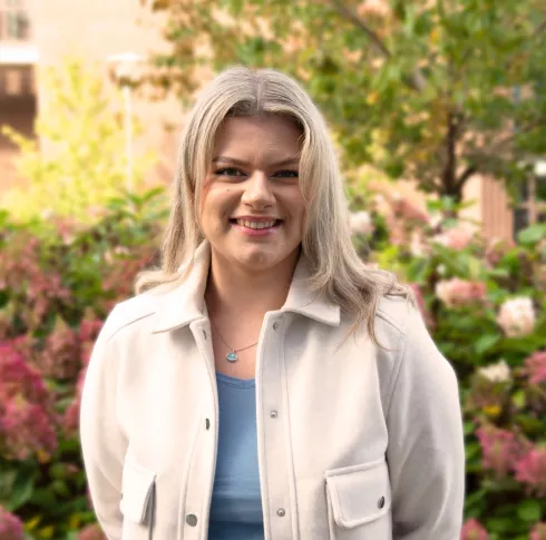 A woman with blonde hair in a beige coat smiling with her arms behind her back