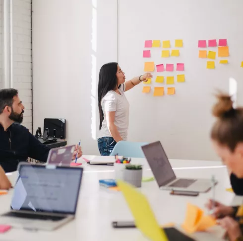 Woman placing sticky notes on a wall as a team of people sitting at a desk with laptops watch her.