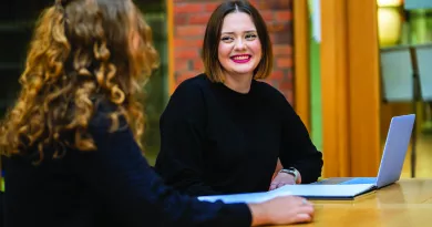 two women sitting at a table, one has her back turned while the other is facing the camera smiling. One of the women has a laptop in front of her