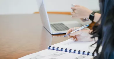 person writing in notebook with another person in background using a laptop