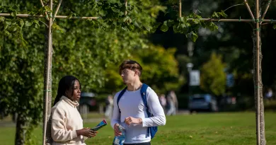 Female student sitting, holding a book, speaking to male student, holding a water bottle.