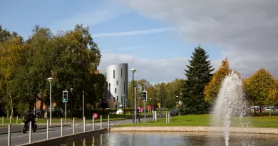 A fountain sprays water into the air. In the background there is a tall silver building slightly covered by trees