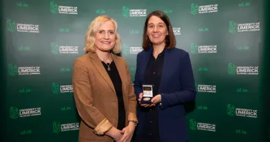 Two women standing in from of a UL branded backdrop. The woman on the right is holding an awards medal 