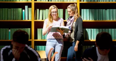 Two female students standing in front of a wall of books looking at a book and two male students sitting at a desk in front of them both reading a book.