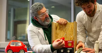 Two men in a lab examining a wooden model with a red toy car and switches.