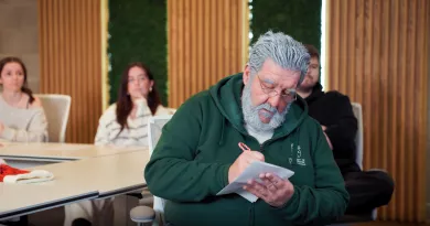 An elderly man in a green hoodie writes in a notepad in a classroom; others sit in the background.