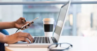 Image depicting a work desk with coffee, glasses, and a laptop. A woman's hand is holding a mobile phone.