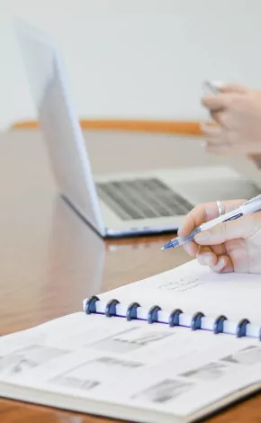 person writing in notebook with another person in background using a laptop