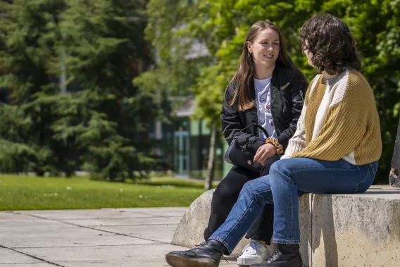 Two girls are sitting outside on a stone bench facing each other chatting. It is a sunny day and there are large trees in the background.