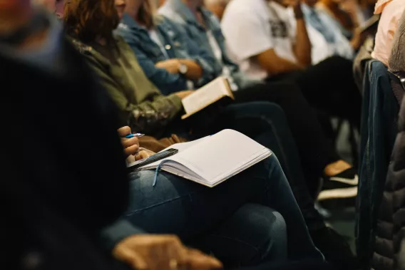 Row of people attending a conferences. They are seated and some rest notebooks on their knees.