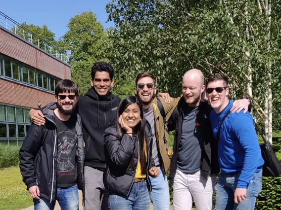 A group of friends, five men and one woman, in casual clothes smiling outside in front of trees