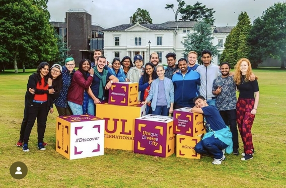 A group of students 20 students standing with colourful boxes with UL written on them, outside on grass