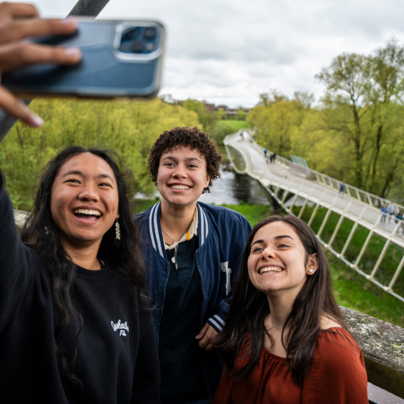 3 students taking a selfie in front of the living bridge on campus