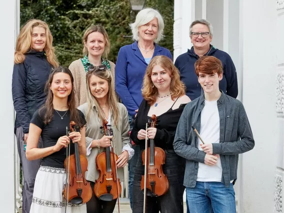 Two rows of eight people smiling at the camera. The bottom row are holding violins.