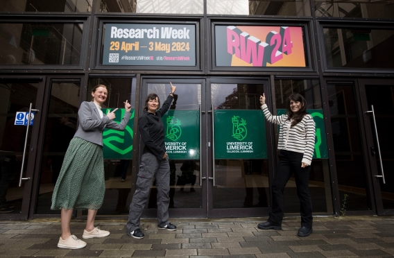 Three people pointing towards a Research Week banner