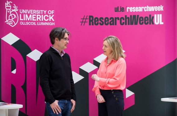 A man and a woman having a conversation in front of a Research Week banner