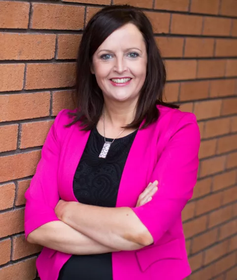 Woman with long brown hair, arms crossed wearing a pink jacket and black dress