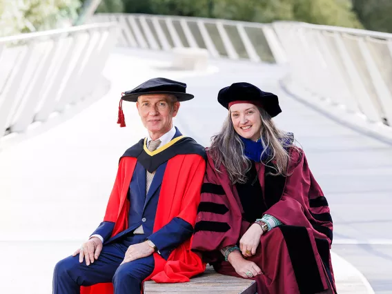 A man in a red graduation gown and a woman in a burgundy graduation gown sitting on a bench smiling at the camera