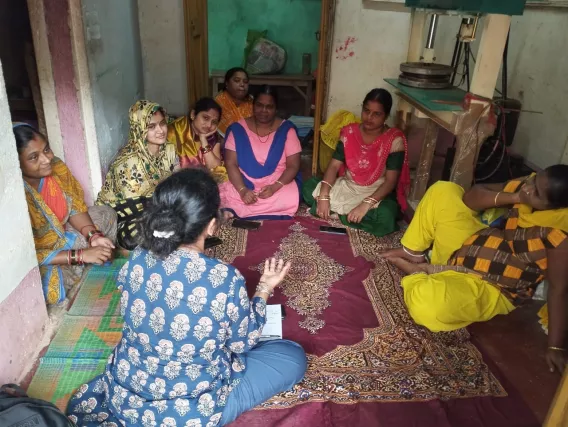 Nita sits across from seven women. They sit on the floor and are having a discussion. 