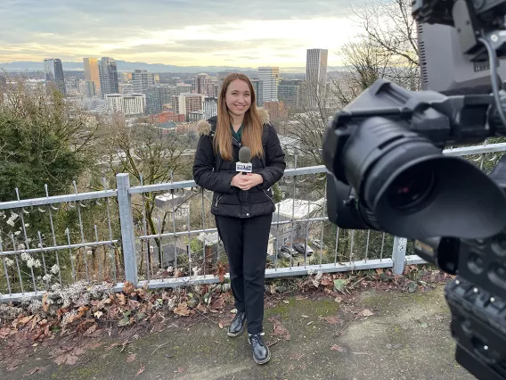 A news reporter holding a microphone talking to the camera standing outside near a fence with a city skyscape in the background