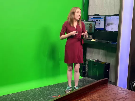 A female reporter in a red dress standing in front of a green screen in a news studio