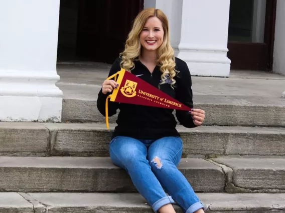 A blonde woman in a black top and jeans sitting on stone steps holding a wine coloured University of Limerick flagth 