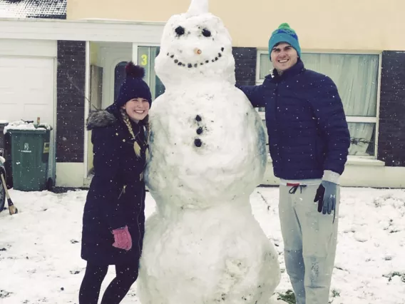 A woman and a man wearing coats and hats standing beside a snowman