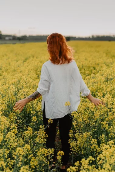 ginger haired girl with tattoo sleeve on left arm in white shirt and black pants walking through field of yellow flowers